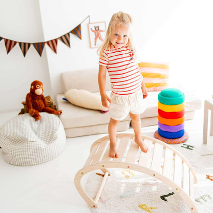 Ein kleines Mädchen steht auf einem Kletterbogen aus Holz in einem bunten Spielzimmer mit Spielzeug.