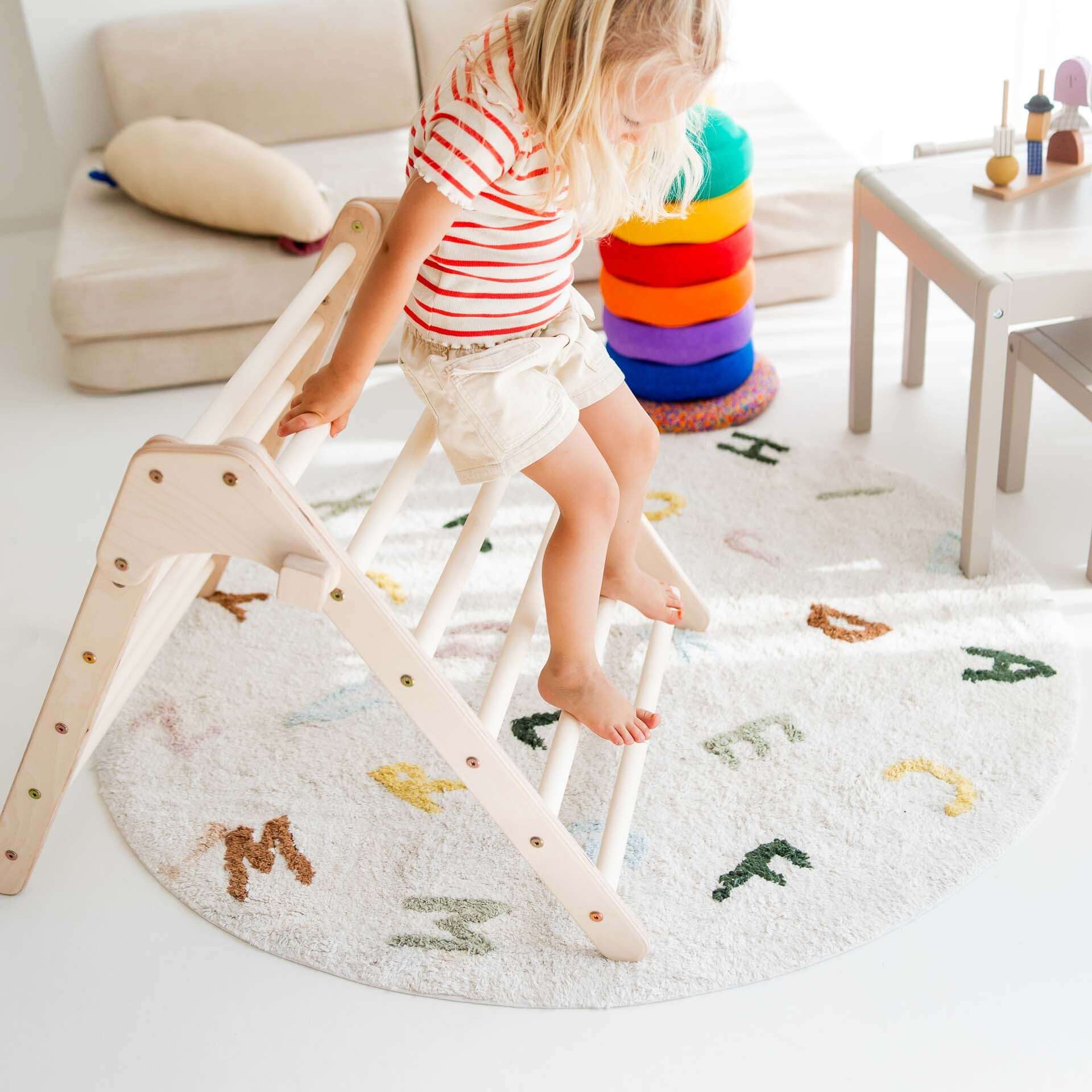 Kind spielt auf dem faltbaren Kletterdreieck Lina aus Holz auf einem bunten Teppich.